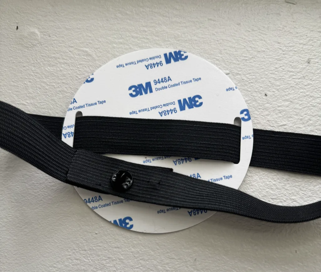 The back of a Tap.Giving plate, showing its 3M adhesive backing, with a black elastic band threaded through the slot on each edge and the two ends joined by a small fastener pin