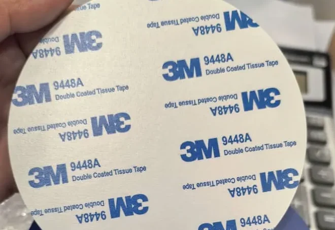 A hand holding a round tap plate face down, showing the 3M 9448A double coated adhesive backing across the whole disc