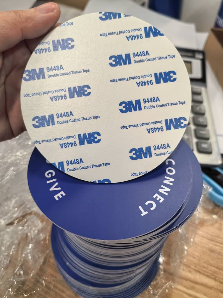 The back of a Tap.Giving plate showing pre-applied 3M 9448A double-coated adhesive tape, held above a stack of navy plates