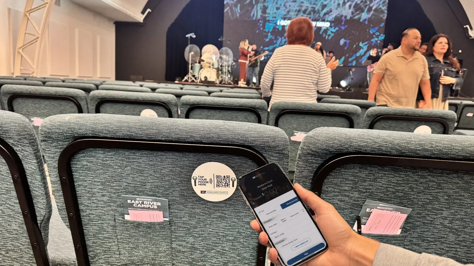 A church auditorium with tap-to-give plates mounted on the chair backs and a congregant tapping a phone to one