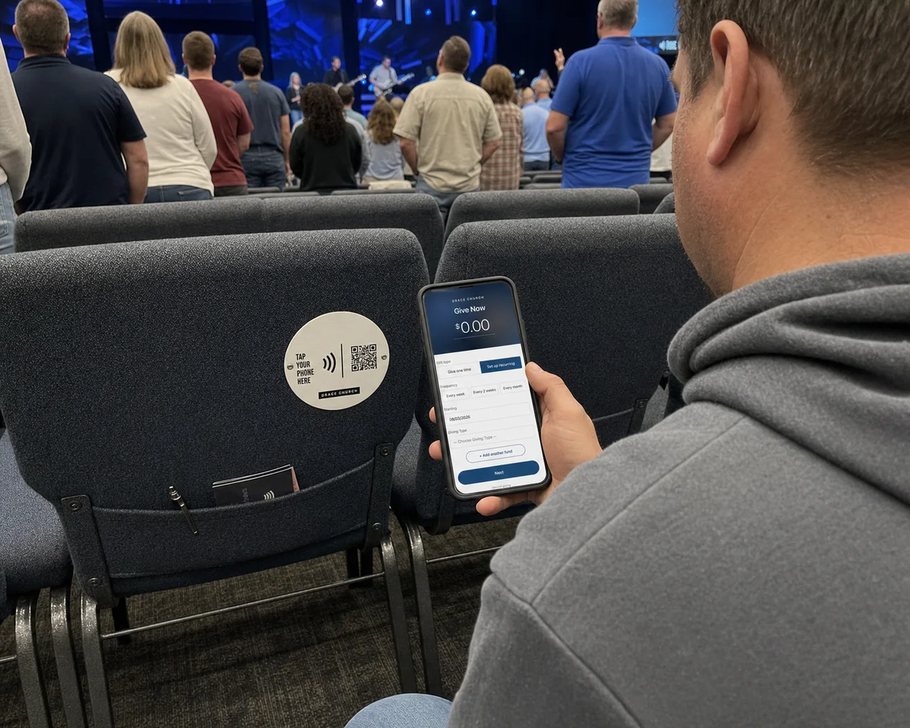 A congregation standing during worship while a seated person taps a phone to the tap-to-give plate on the chair back in front of them