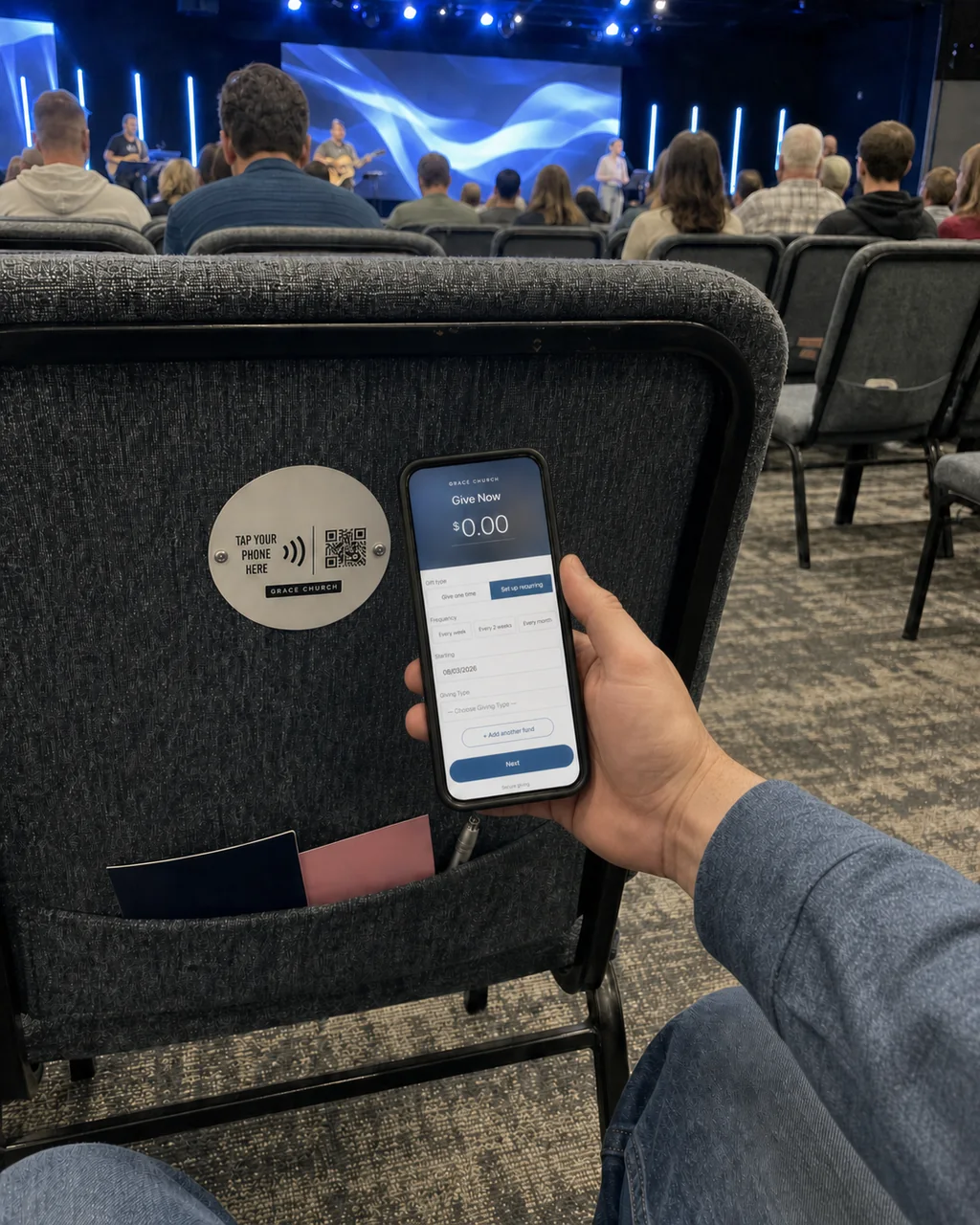 A hand holding a phone to a tap plate mounted on the back of a chair, with the church's giving page open on screen.